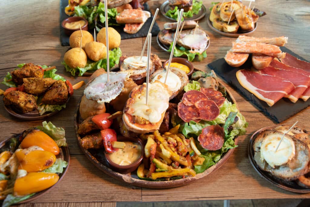 Grande planche de tapas variés, incluant jambon cru, croquettes, toasts au fromage de chèvre, légumes grillés et poulet sur table en bois.